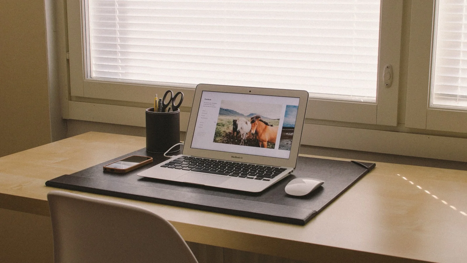 An open laptop on a tidy light wooden desk beside a window with soft blinds, in calm natural light, with a pen holder and a phone nearby.