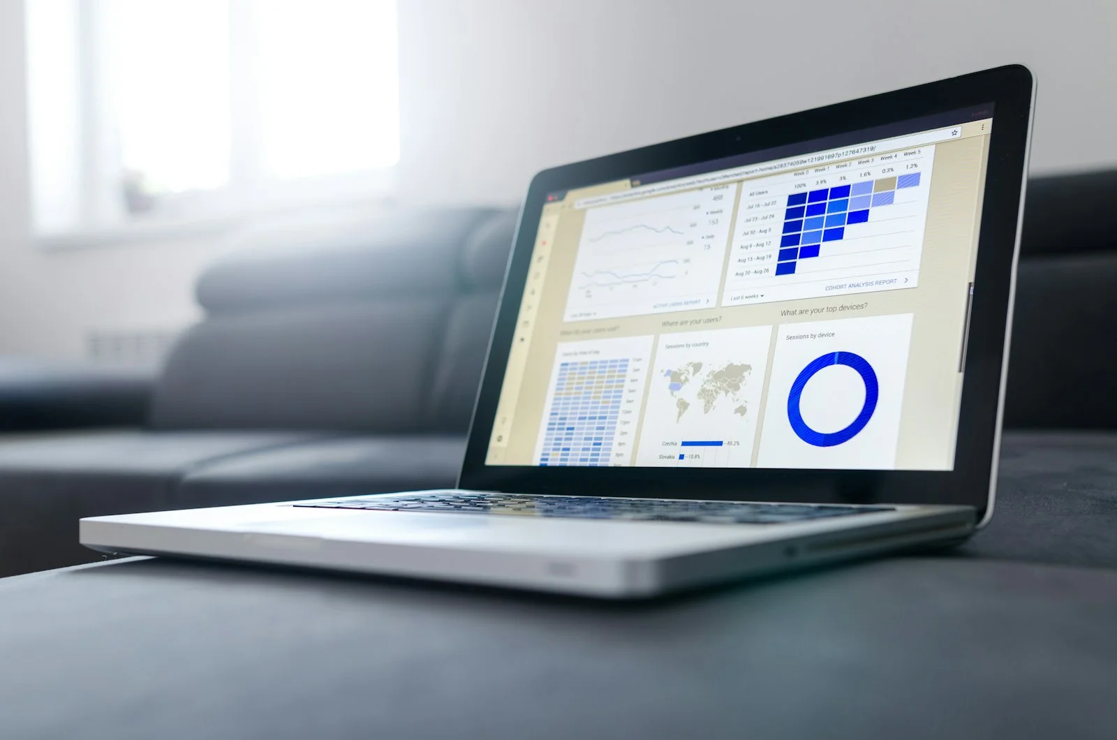 A laptop on a table in a bright, calm living room, its screen showing a data dashboard with a heatmap grid, a world map and a blue donut chart. No people or logos are visible.