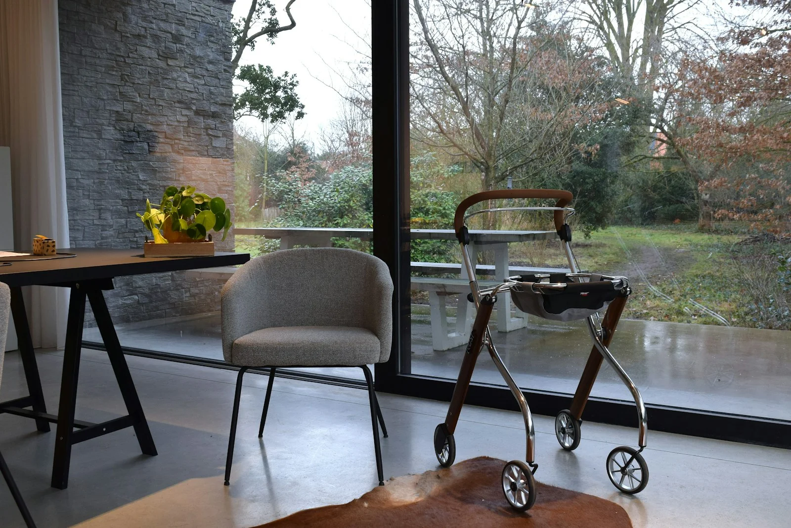 A rollator walking frame and an upholstered chair beside a large window looking out to a quiet garden, in a calm, naturally lit room.