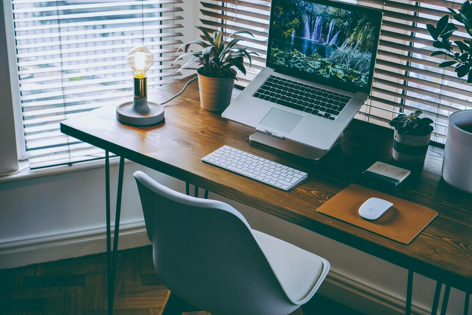 A quiet modern workstation by a window in natural light: a wooden desk, an open laptop, a keyboard and a small plant, with no people and no readable text.